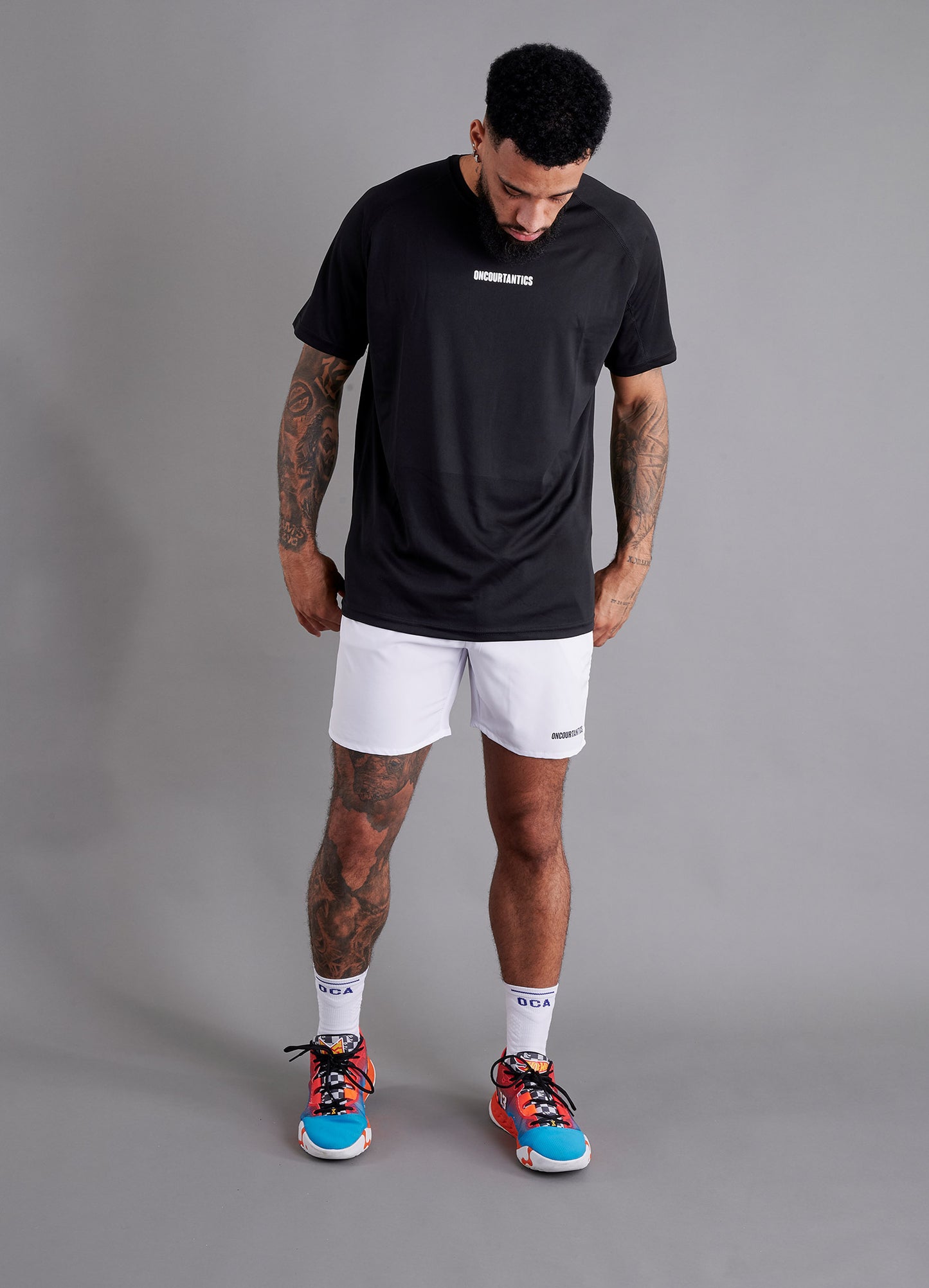 Male basketball model wearing ONCOURTANTICS black performance t-shirt with small chest logo, paired with white shorts, front-facing against plain grey background.