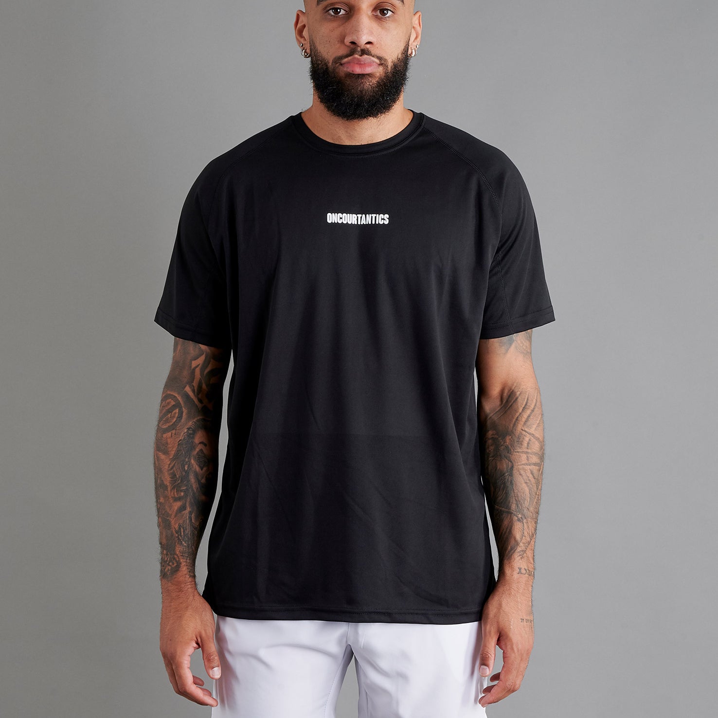Male basketball model wearing ONCOURTANTICS black performance t-shirt with small chest logo, paired with white shorts, front-facing against plain grey background.