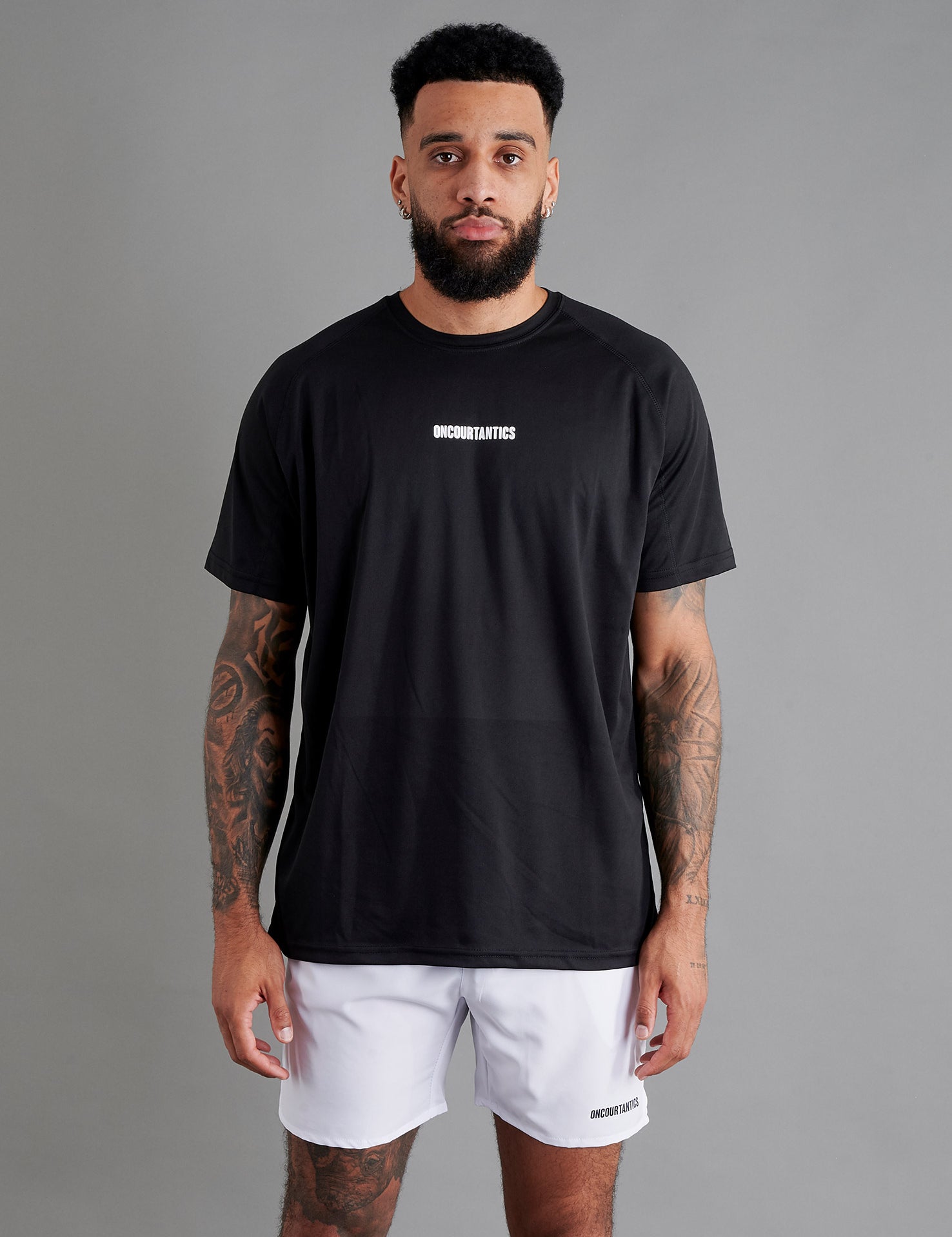 Male basketball model wearing ONCOURTANTICS black performance t-shirt with small chest logo, paired with white shorts, front-facing against plain grey background.