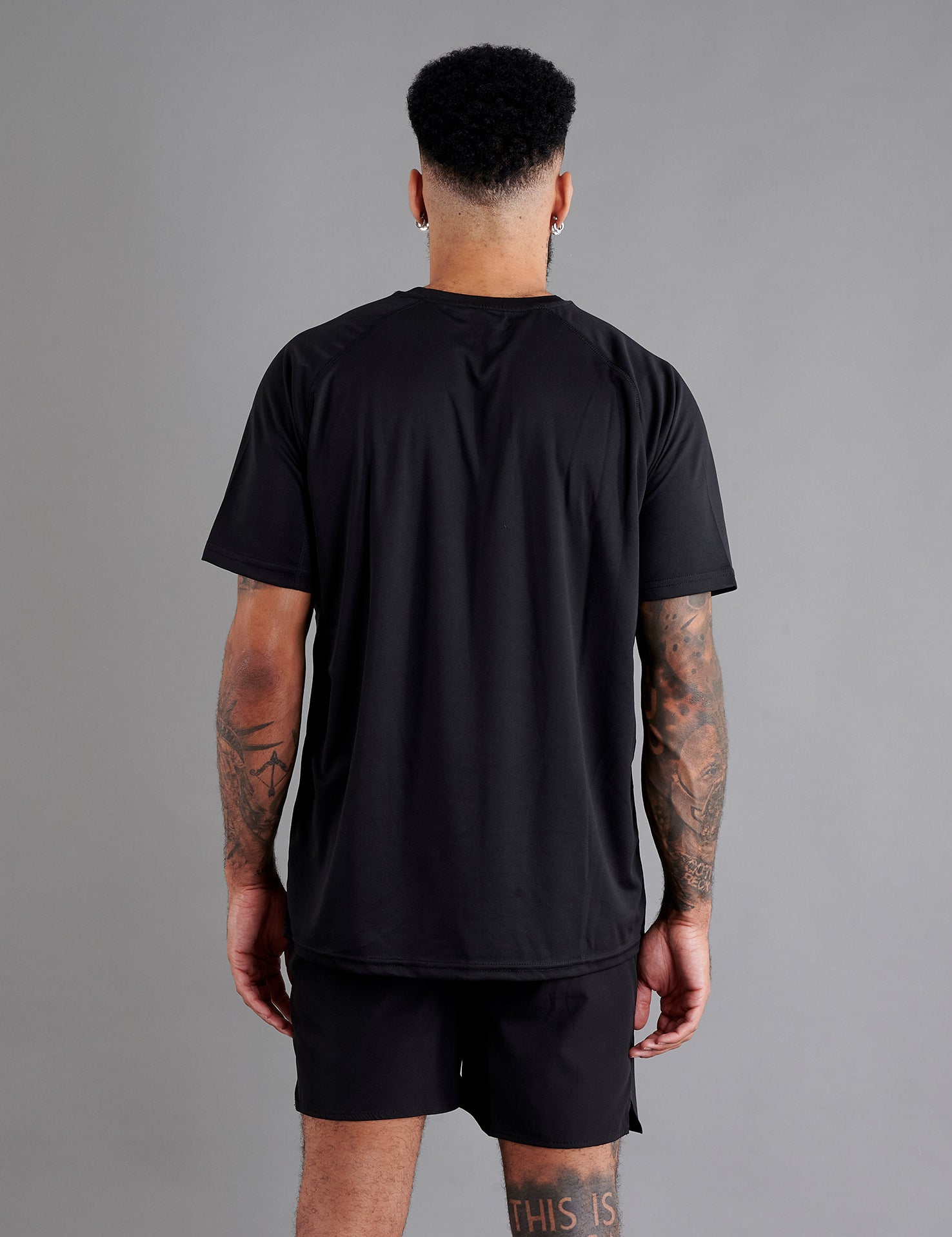 Rear view of male model in ONCOURTANTICS black performance t-shirt with small chest logo, white shorts, standing on grey background.