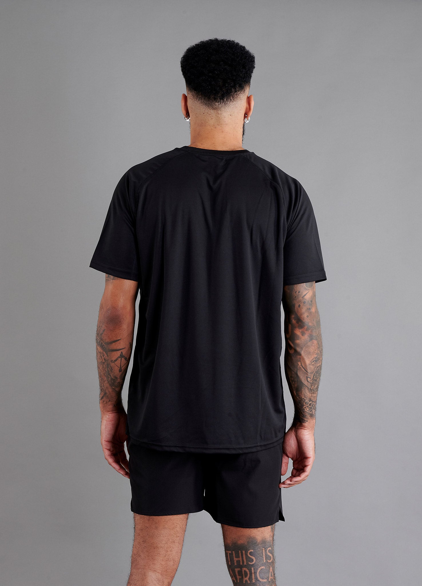 Rear view of male model in ONCOURTANTICS black performance t-shirt with small chest logo, white shorts, standing on grey background.