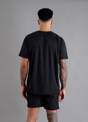 Rear view of male model in ONCOURTANTICS black performance t-shirt with small chest logo, white shorts, standing on grey background.