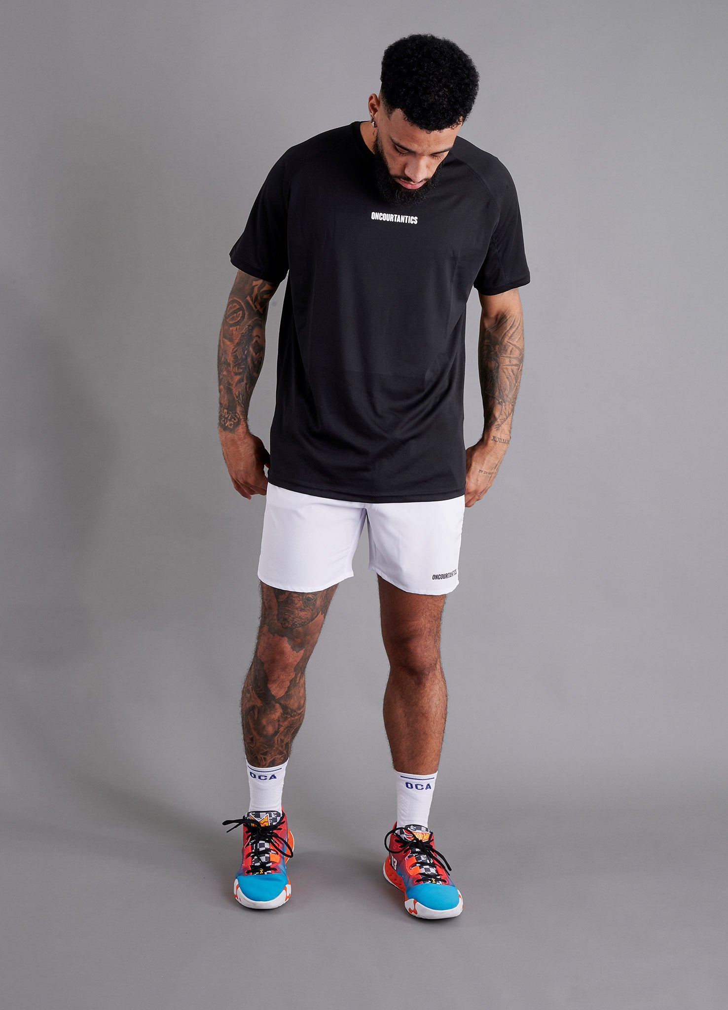 Male basketball model wearing ONCOURTANTICS black performance t-shirt with small chest logo, paired with white shorts, front-facing against plain grey background.