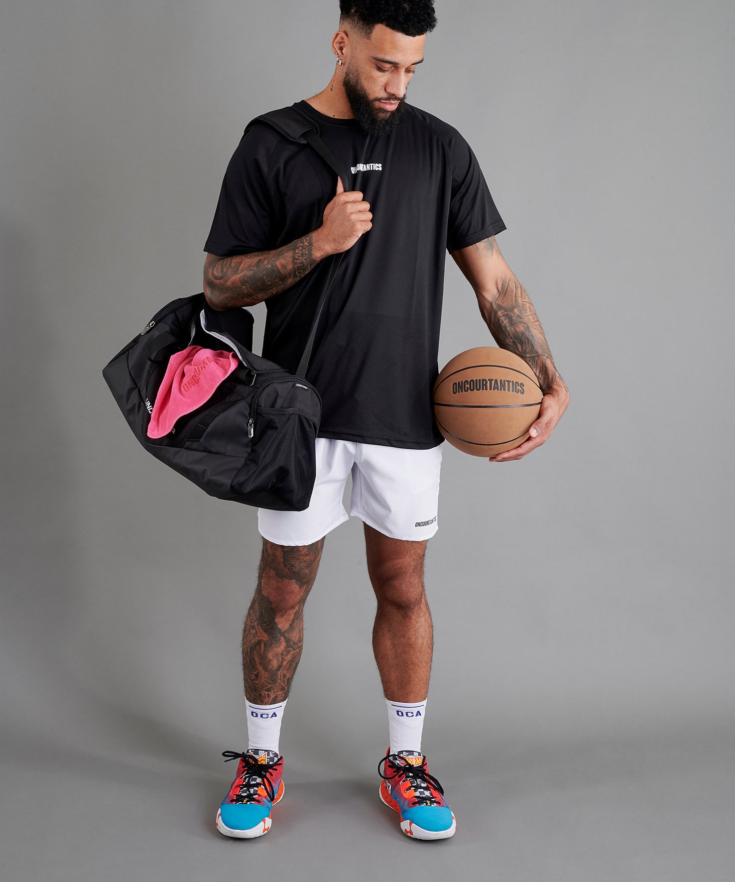 Male basketball model wearing ONCOURTANTICS black performance t-shirt with small chest logo, paired with white shorts, front-facing against plain grey background, holding a holdall sports back, with a pink towel and an oncourtantics basketball.