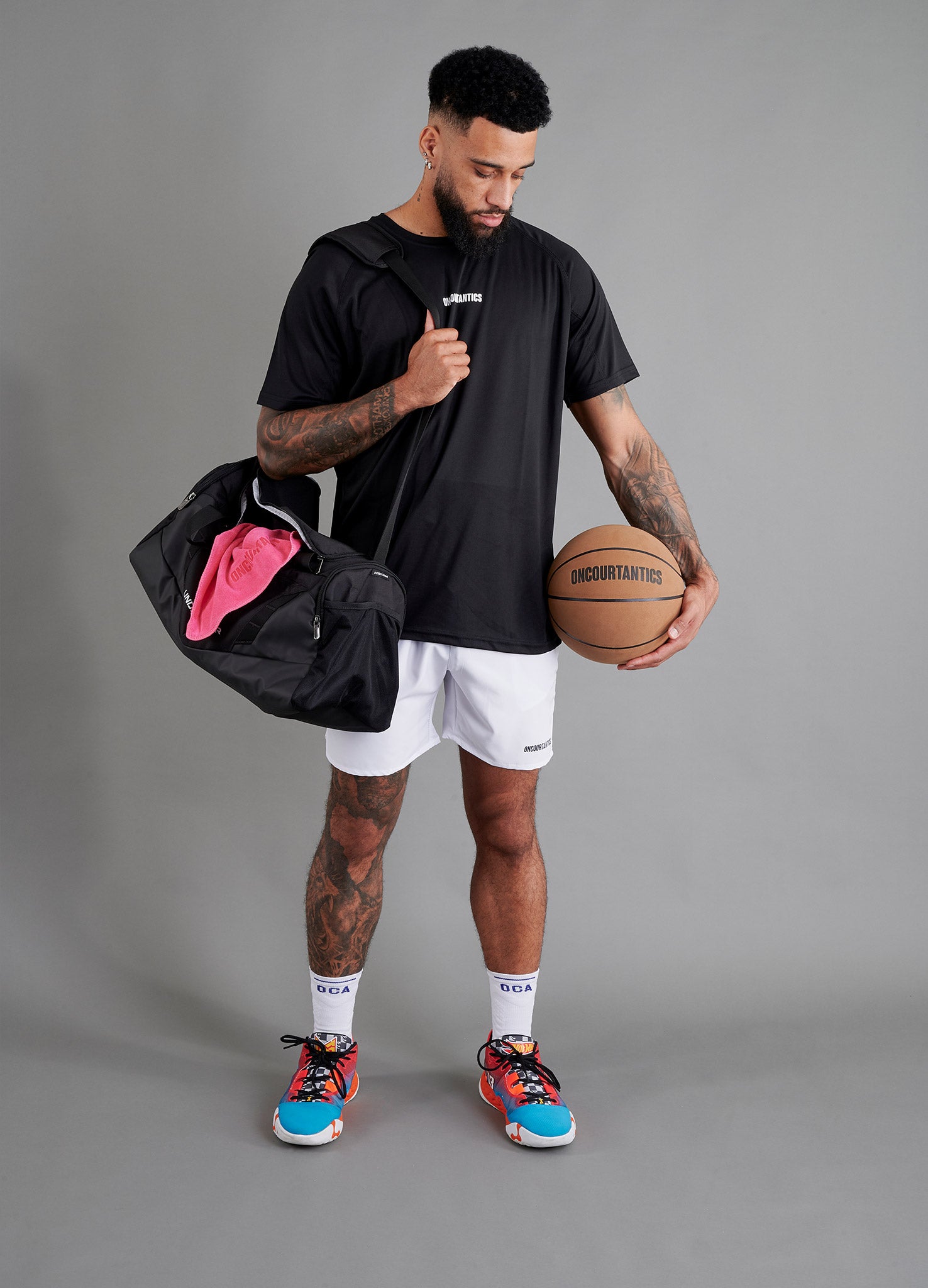 Male basketball model wearing ONCOURTANTICS black performance t-shirt with small chest logo, paired with white shorts, front-facing against plain grey background, holding a holdall sports back, with a pink towel and an oncourtantics basketball.