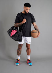 Male basketball model wearing ONCOURTANTICS black performance t-shirt with small chest logo, paired with white shorts, front-facing against plain grey background, holding a holdall sports back, with a pink towel and an oncourtantics basketball.