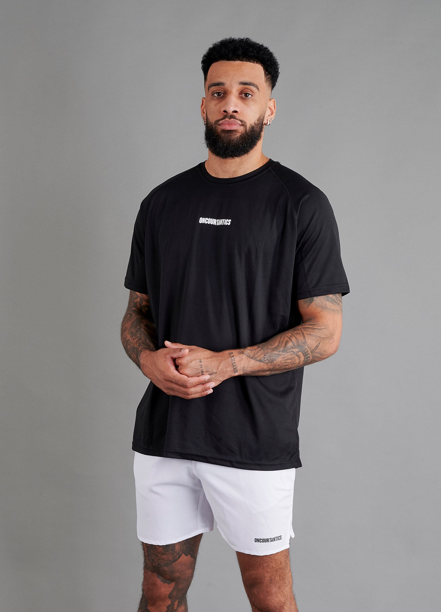 Male basketball model wearing ONCOURTANTICS black performance t-shirt with small chest logo, paired with white shorts, front-facing against plain grey background.