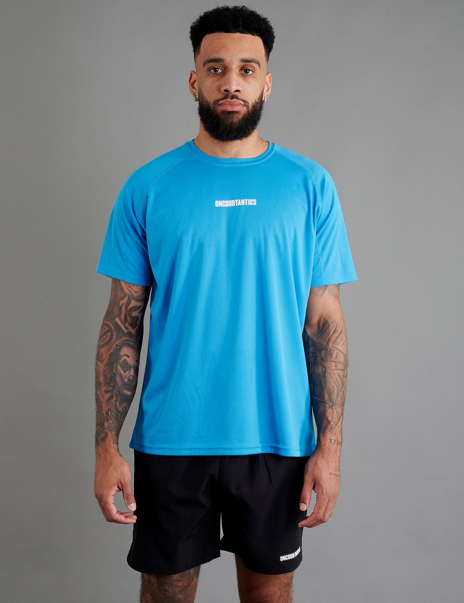 Male basketball model wearing ONCOURTANTICS blue performance t-shirt with small chest logo, paired with black shorts, front-facing against plain grey background.