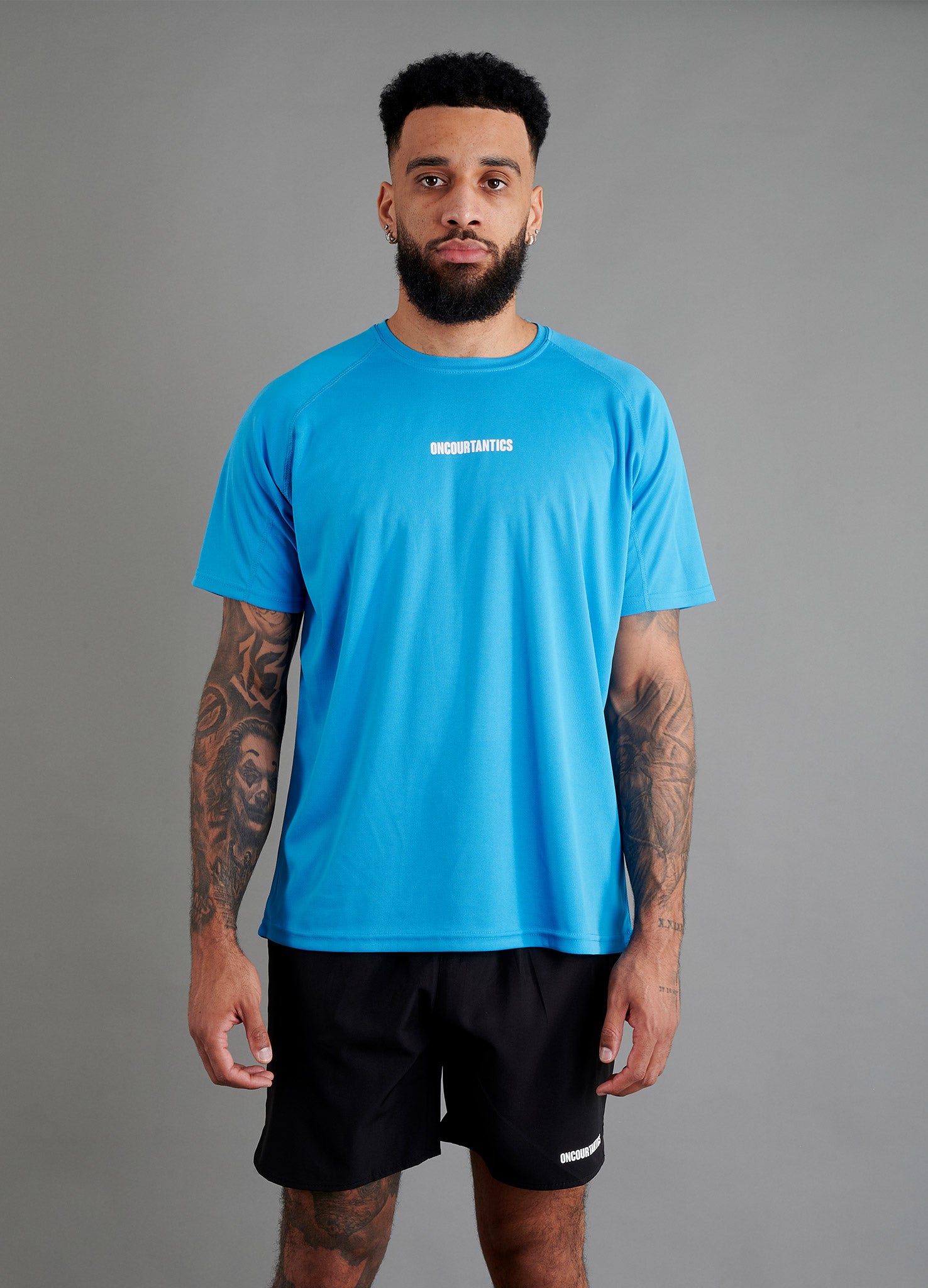 Male basketball model wearing ONCOURTANTICS blue performance t-shirt with small chest logo, paired with black shorts, front-facing against plain grey background.