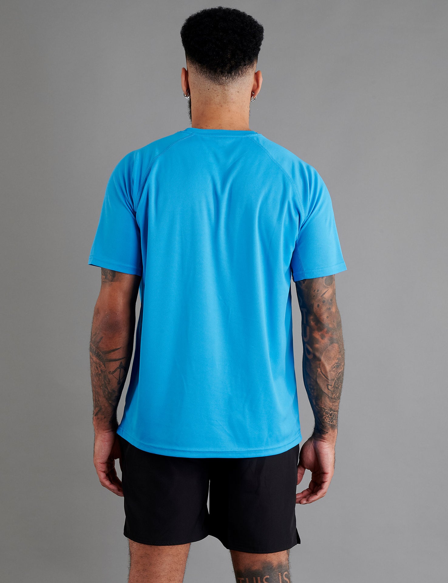 Rear view of male model in ONCOURTANTICS blue performance t-shirt with small chest logo, black shorts, standing on grey background