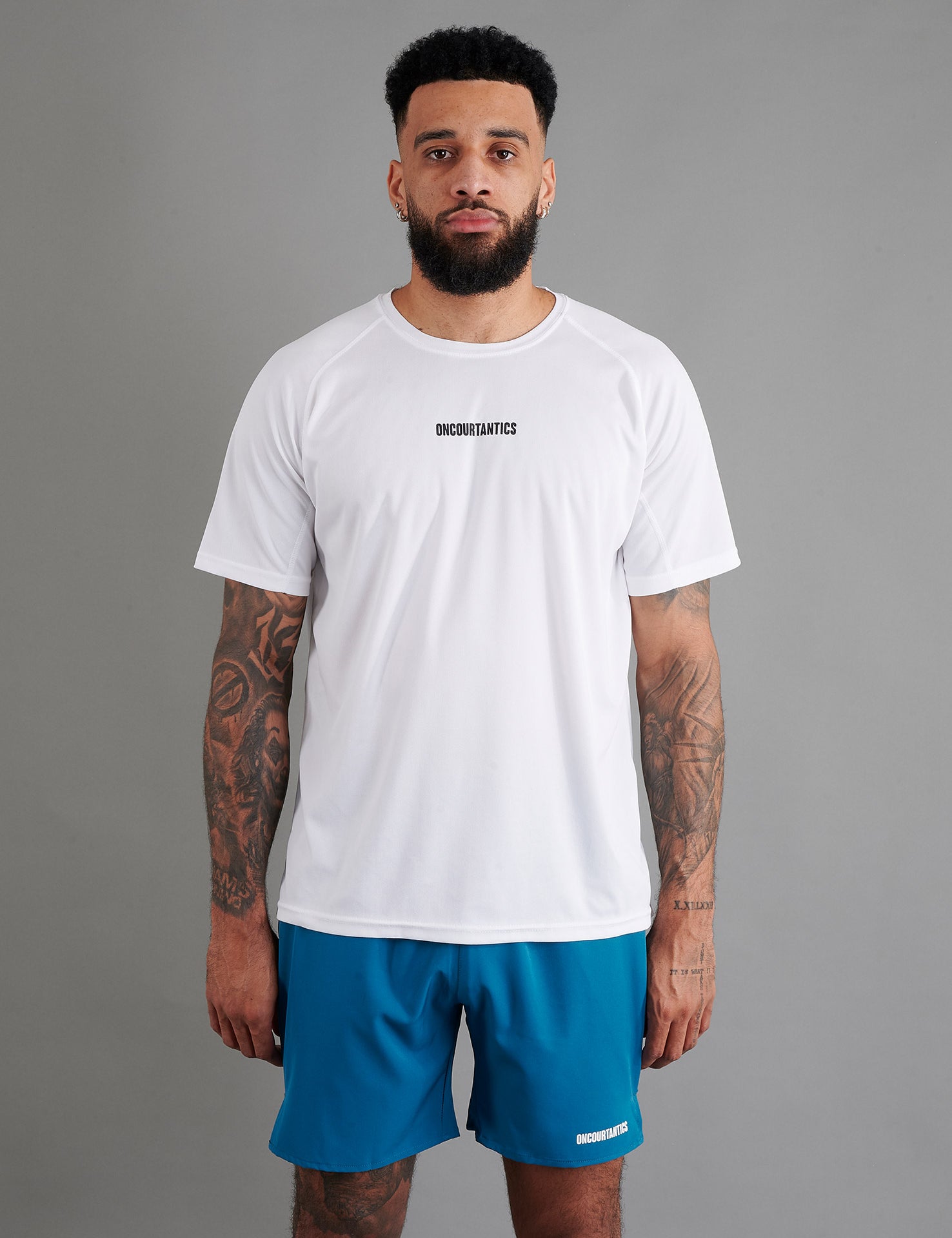 Male basketball model wearing ONCOURTANTICS White performance t-shirt with small chest logo, paired with teal shorts, front-facing against plain grey background.