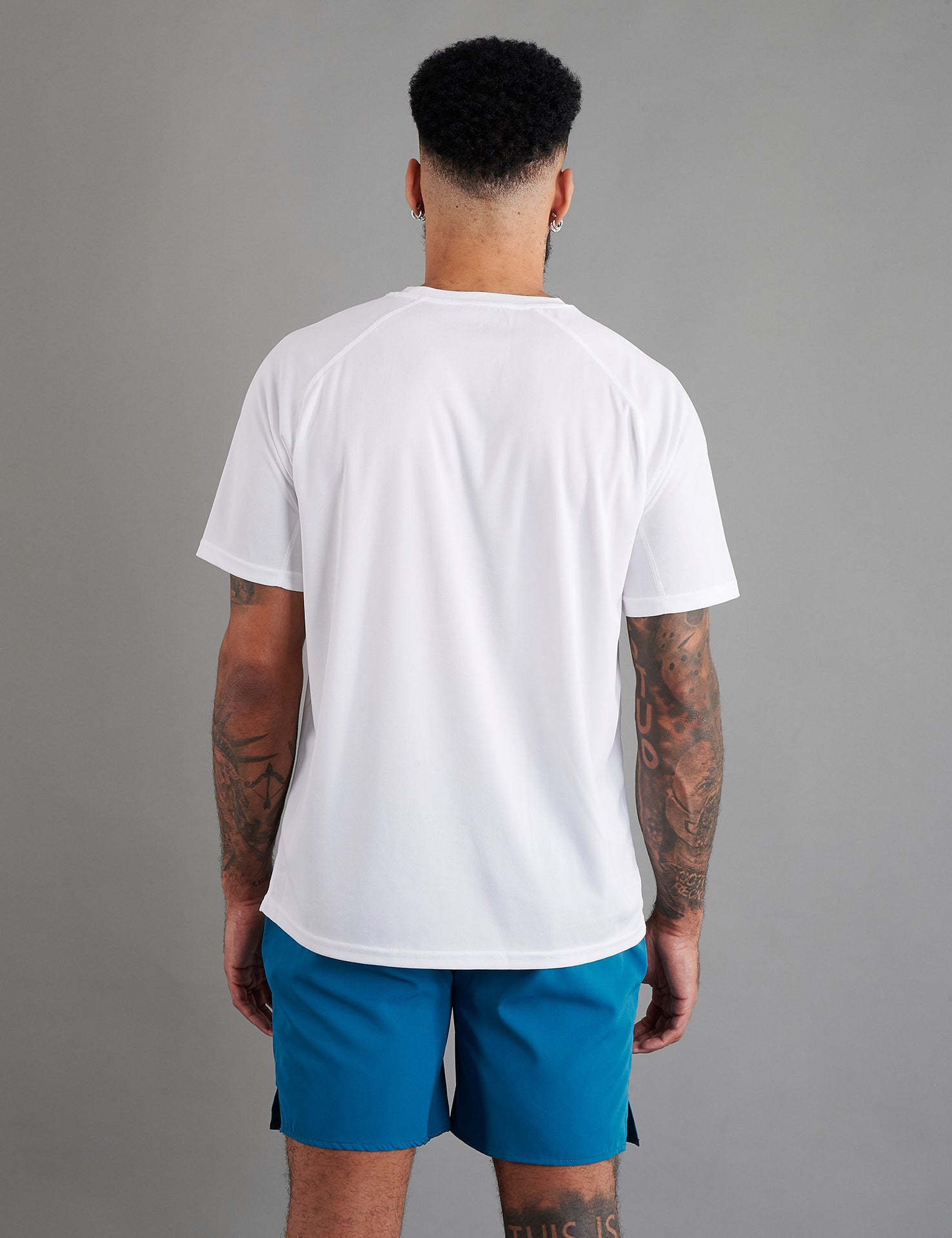 Rear view of male model in ONCOURTANTICS white performance t-shirt with small chest logo, teal shorts, standing on grey background.