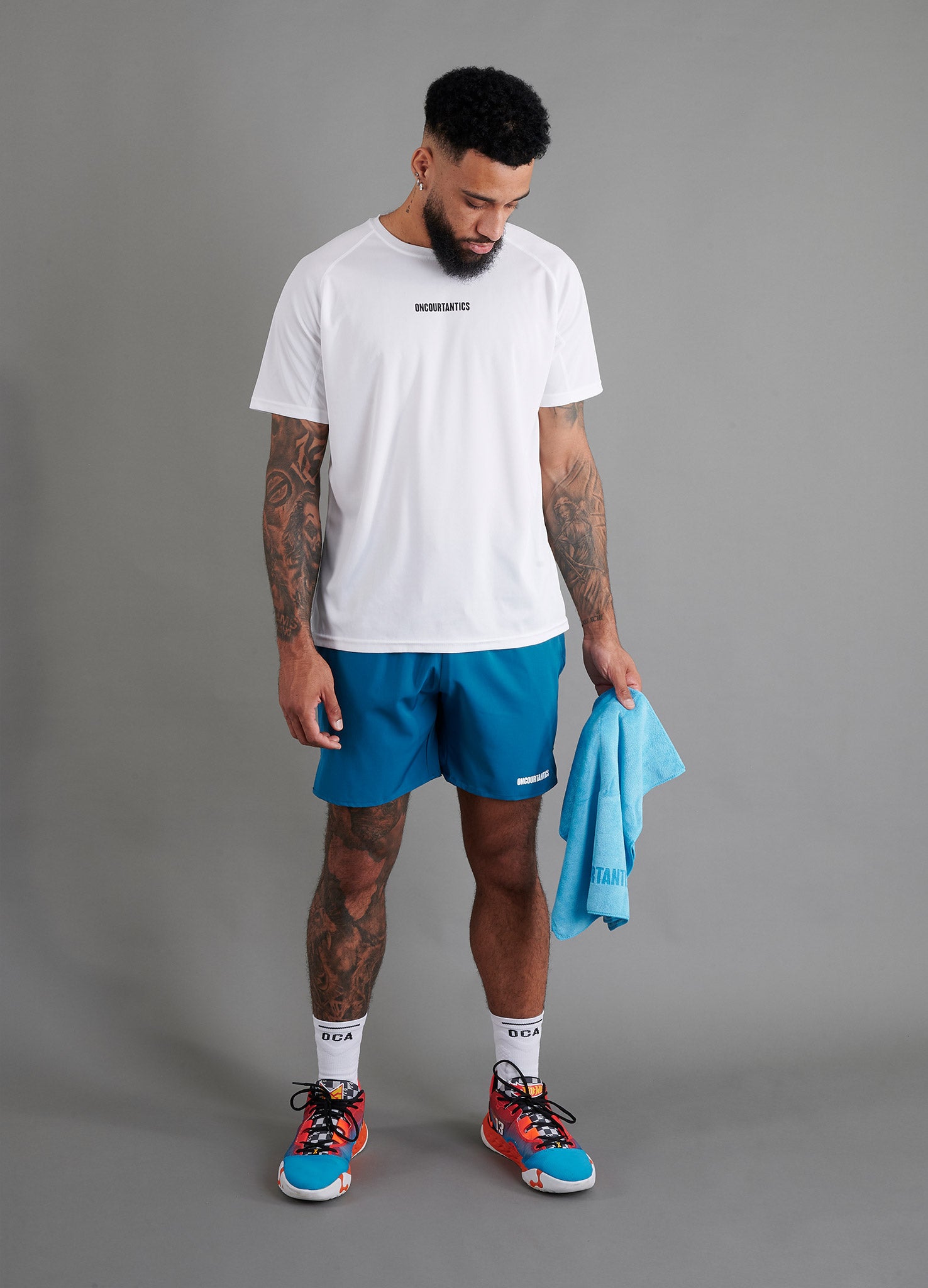 Male basketball model wearing ONCOURTANTICS black performance t-shirt with small chest logo, paired with teal shorts, holding a blue microfibre towel in hand, front-facing against plain grey background